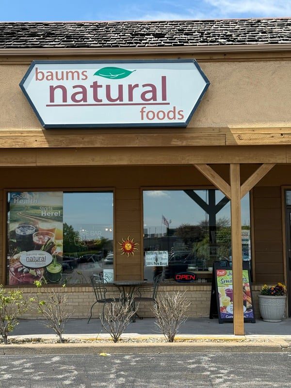 15c63163d816769bb85f2f471e16648a Storefront of Baums Natural Foods in Highland, IN, featuring a large sign with the store name and logo, promotional posters for fresh juices and smoothies, outdoor seating with metal chairs and a table, reflecting a local natural health food store offering vitamins, supplements, holistic products, and wellness services.