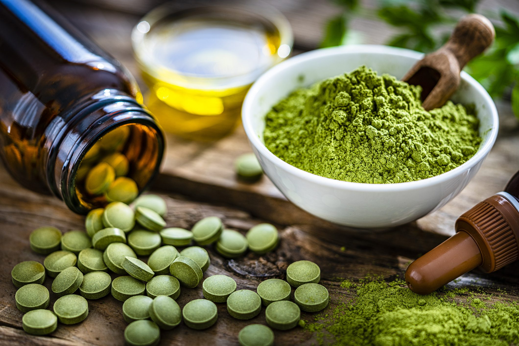 Moringa oleifera powder and pills on rustic table Green herbal supplement tablets spilling from amber bottle next to bowl of green powder with wooden scoop and dropper bottle on rustic wooden surface, representing natural vitamins and holistic health products.