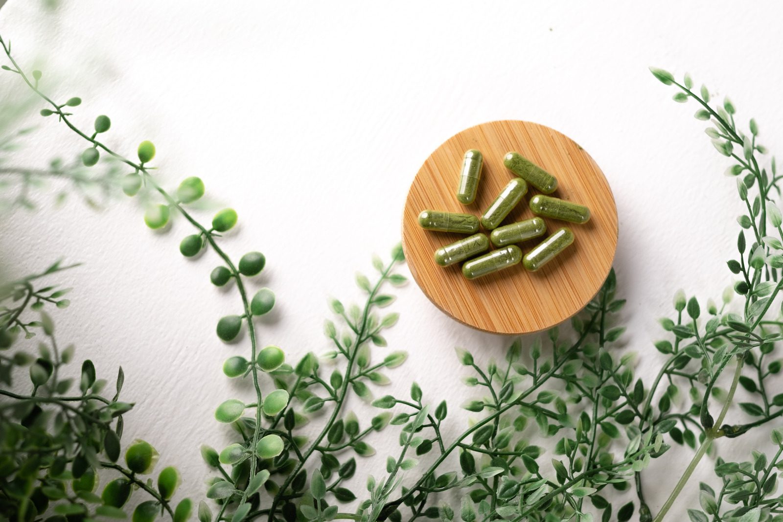 b702d2d8d1e2fabb1663fdd5d759fa83 Green herbal supplement capsules arranged on a round bamboo plate surrounded by green leafy branches on a white background, representing natural vitamins and holistic health products.
